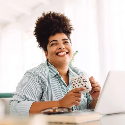 Smiling woman with curly hair holding a coffee mug while sitting at a desk with a laptop in bright room.