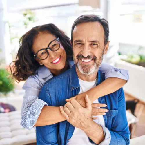 Smiling middle-aged couple hugging happily at home with natural light and cozy background.