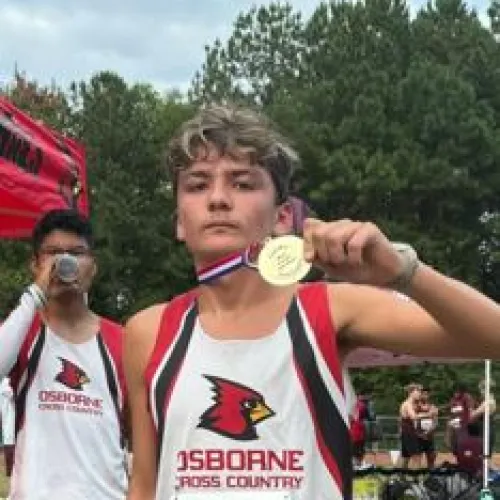 Osborne cross country runner proudly displays gold medal after race with teammate in background outdoors.