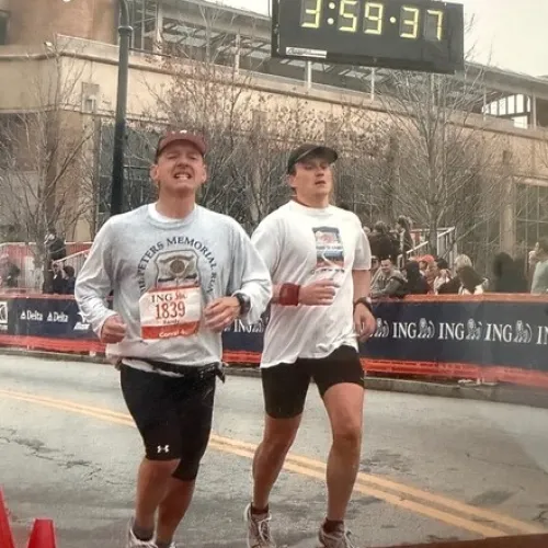Two male runners crossing a marathon finish line, one wearing a race bib and sweatshirt, the other in shorts and a hat.