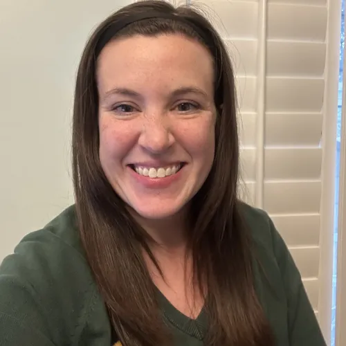 Smiling woman with straight brown hair wearing a green sweater and black headband in front of white shutters