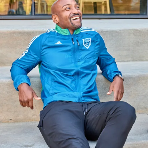 Smiling man in blue athletic jacket and black pants sitting on outdoor concrete steps relaxed.