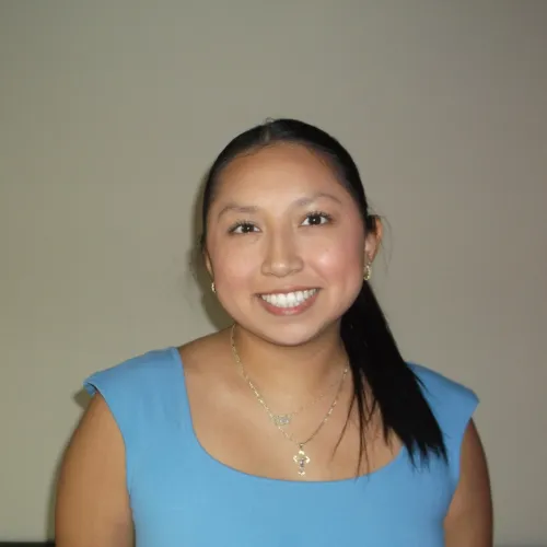 Smiling young woman with long black hair wearing a blue top and gold jewelry against a plain background