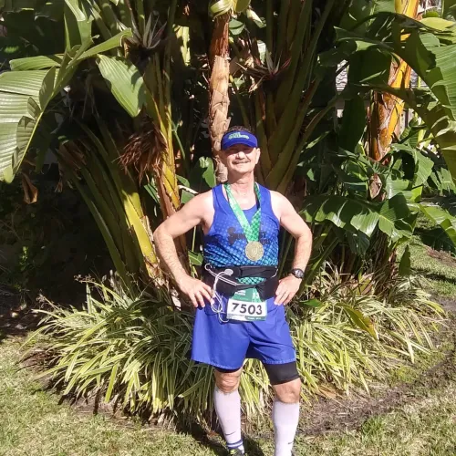 Smiling male runner wearing medal and race bib 7503 poses in blue athletic gear by tropical plants on sunny day