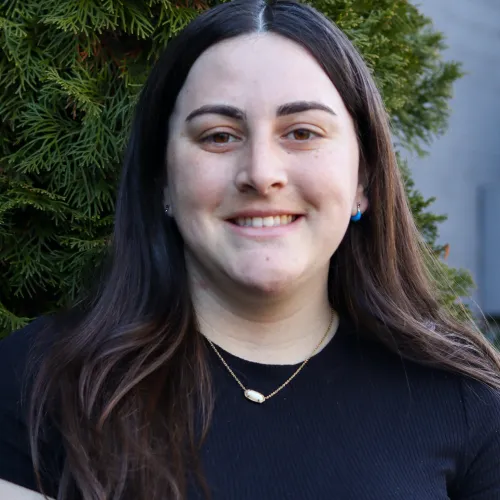 Smiling young woman with long dark hair wearing a black shirt and necklace standing outdoors by green foliage.