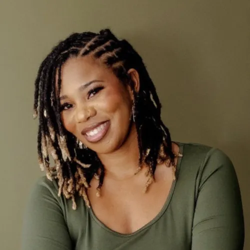 Smiling woman with dreadlocks and hoop earrings wearing a green top against an olive background.