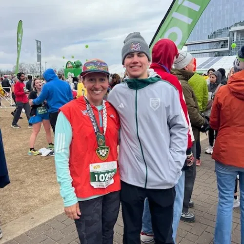 Two smiling young runners wearing race bibs and medals stand together after a marathon event with a green finish line banner in the background