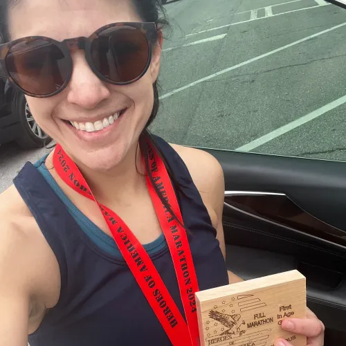Smiling woman wearing sunglasses and marathon medal holding Heroes of America Marathon plaque after race.