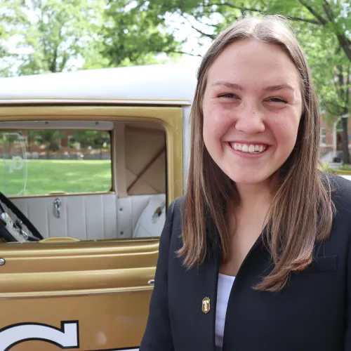 Smiling young woman in black blazer stands beside vintage gold and white car outdoors with green trees in background