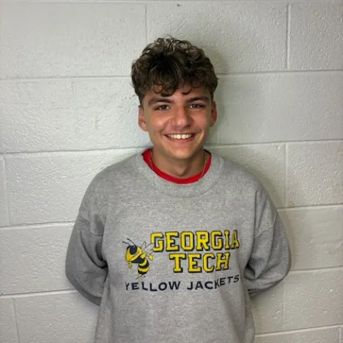 Smiling young man wearing a gray Georgia Tech Yellow Jackets sweatshirt standing against a white brick wall.