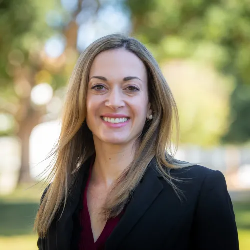 Smiling woman with long blonde hair wearing a black blazer outdoors with blurred green trees in background