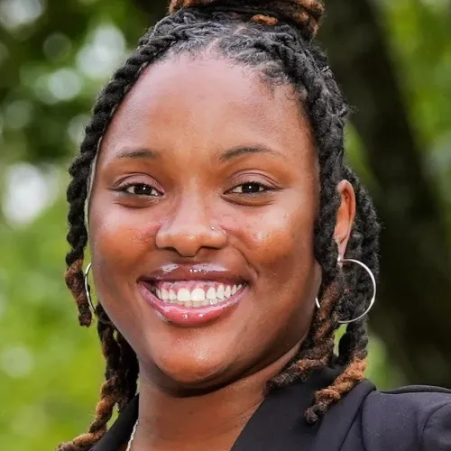 Smiling woman with braided hair and hoop earrings wearing a black outfit outdoors with blurred green background.