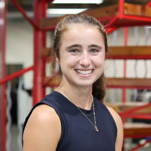 Smiling young woman with a ponytail wearing a black tank top and necklace indoors, staircase background.