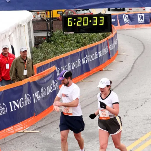 Two runners cross the Georgia Marathon finish line with a digital clock showing 6:20:33 under a blue banner.