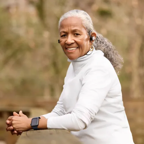 Smiling elderly woman in white sportswear with wireless earphones and smartwatch enjoying outdoor activity.