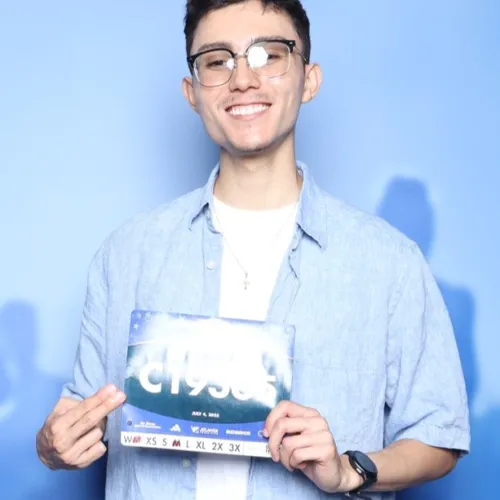 Young man smiling and holding a race bib at an AJC Peachtree Road Race event backdrop with runners behind.