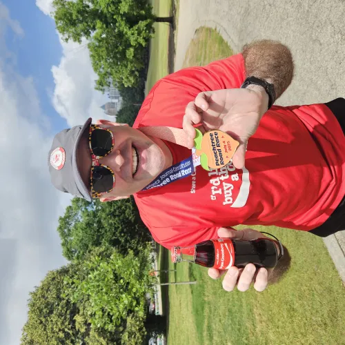 Smiling man in red shirt and cap holds a medal and Coca-Cola bottle outdoors on a sunny day.