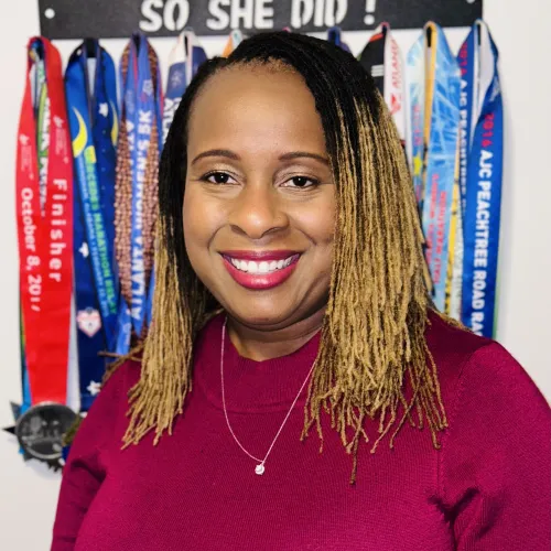 Smiling woman with blonde dreadlocks in maroon top stands in front of race medal display and motivational quote.