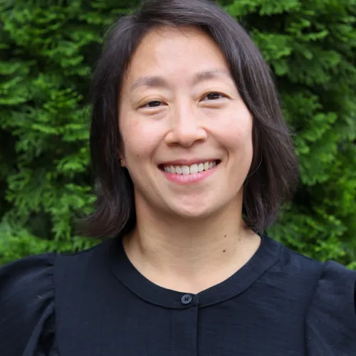 Smiling woman with short dark hair wearing a black buttoned blouse standing outdoors with green foliage background