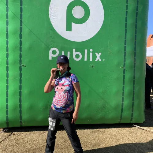Runner in colorful shirt bites medal standing in front of large green Publix banner outdoors on grass.