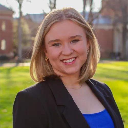 Smiling young woman with blonde hair wearing a black blazer and blue top standing outdoors on a sunny day