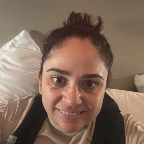 Woman with dark hair in a bun wearing a beige T-shirt and hydration vest, smiling indoors on a bed.