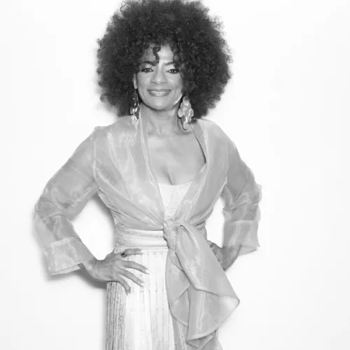 Woman with voluminous curly hair wearing sheer top and large earrings smiling confidently with hands on hips.