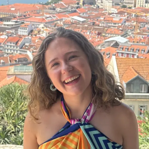 Young woman smiling on a stone terrace overlooking a city with red rooftops and a river under a clear blue sky.