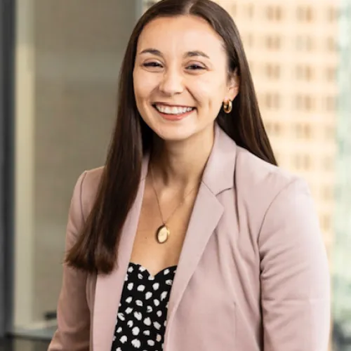 Smiling woman with long brown hair wearing a pink blazer and black patterned top in office setting.