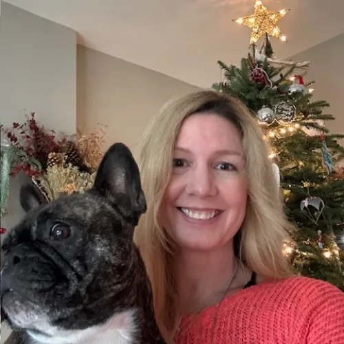 Smiling woman with blonde hair holds a brindle French bulldog in front of a decorated Christmas tree with a glowing star topper.