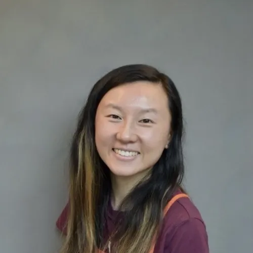 Smiling young woman with long dark hair wearing a maroon shirt and orange apron against a gray background