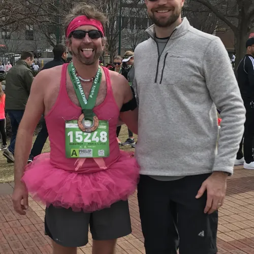 Two men posing at a marathon event, one wearing a pink tutu and race bib, the other in casual sportswear and a red cap.