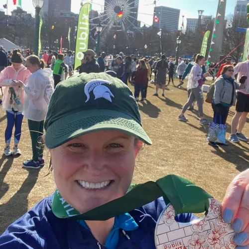 Smiling woman in a green cap holding a finisher medal at an outdoor event with a ferris wheel and crowd in the background.