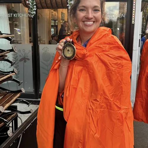 Smiling woman wrapped in orange blanket holding a race finisher medal outside a building with flower decorations.