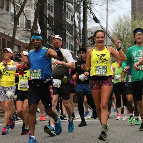 Diverse group of runners smiling and gesturing peace signs during an outdoor city marathon event.