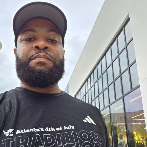 Man with beard wearing black Atlanta's 4th of July Tradition T-shirt and gray cap outside a modern building.