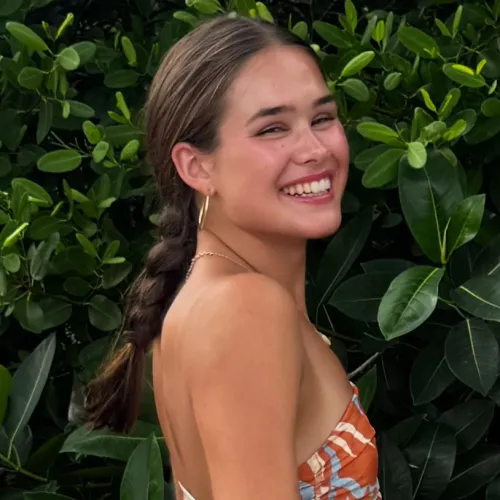 Smiling woman in a patterned dress with braided hair standing in front of lush green foliage outdoors.