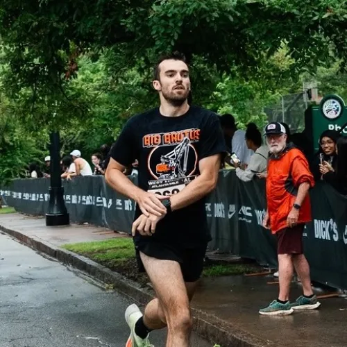 Male runner in black shorts and shirt crossing a street checkpoint with spectators in the background.