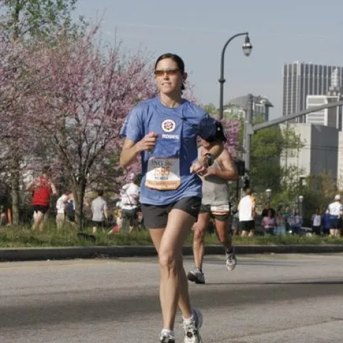 Female runner in blue shirt and sunglasses competing in an outdoor city marathon with other runners and blossoming trees.