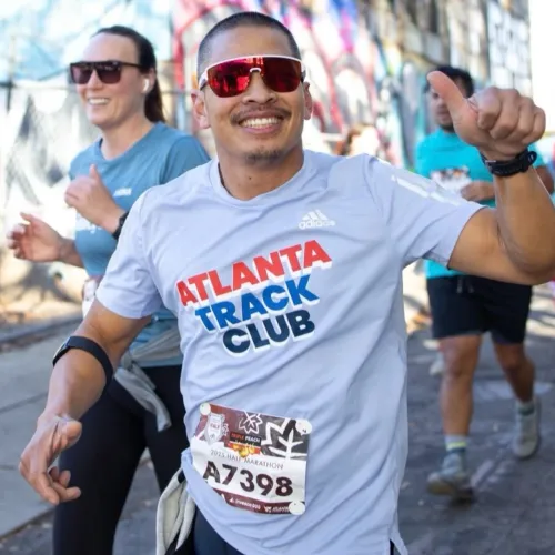 Smiling male runner wearing Atlanta Track Club shirt and race bib giving thumbs up during outdoor marathon.