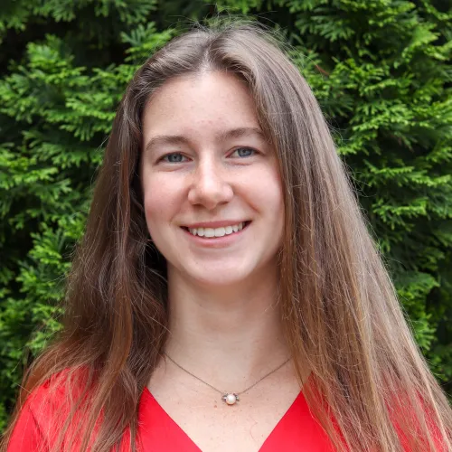 Young woman with long brown hair smiling outdoors in front of green foliage, wearing a red top and pearl necklace