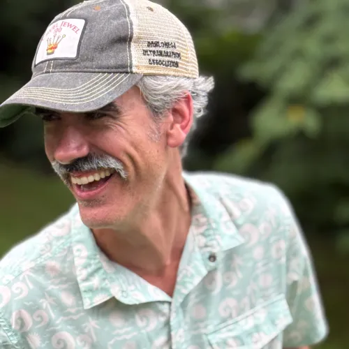 Smiling man with gray mustache wearing a patterned light green shirt and trucker hat outdoors with blurred greenery.