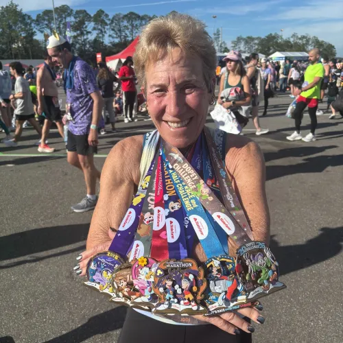 Smiling senior woman holding multiple colorful Disney marathon medals at outdoor race event on sunny day