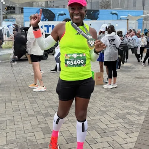 Smiling female runner in neon green shirt and red visor holding a medal celebrating marathon finish outdoors.