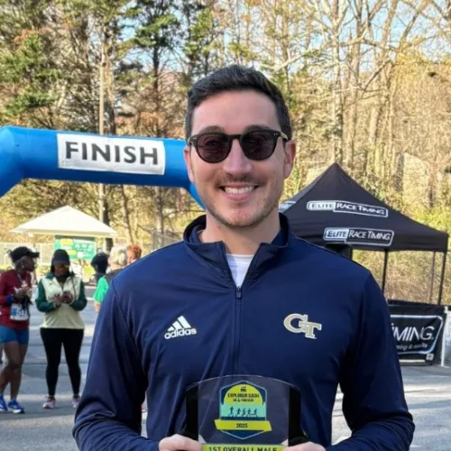 Male runner wearing sunglasses and neon green shoes holds a marathon award at outdoor finish line event on sunny day.