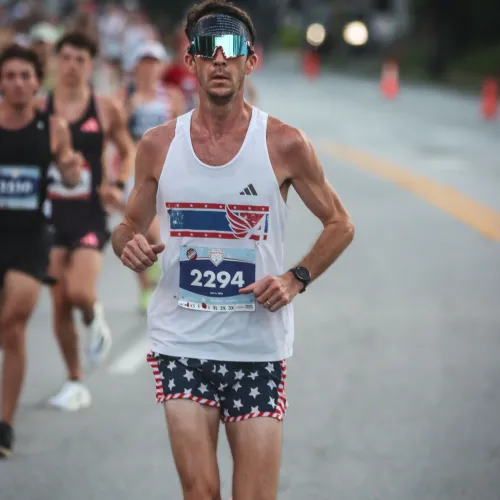 Male runner wearing reflective sunglasses and star-spangled shorts competing in a road race with others behind him.