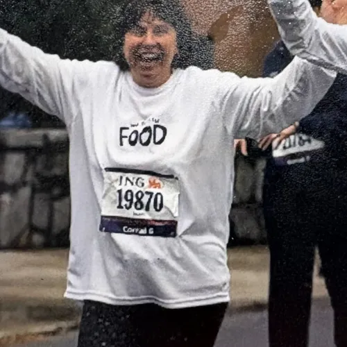 Smiling woman wearing a white shirt and race bib 19870 with arms raised during an outdoor run or race.