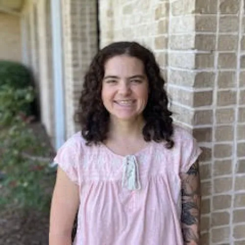 Smiling woman with curly hair wearing a pink blouse standing by a brick wall with greenery nearby.