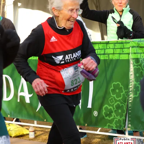 Elderly woman smiling and running in Atlanta Marathon with supportive spectator cheering behind green banner