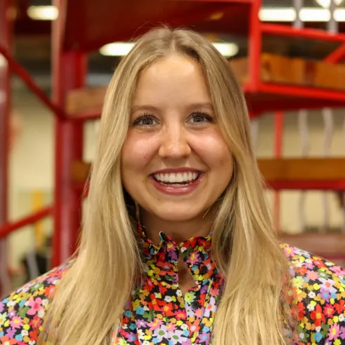 Smiling young woman with long blonde hair wearing a colorful floral blouse in an indoor setting with red stair railings.